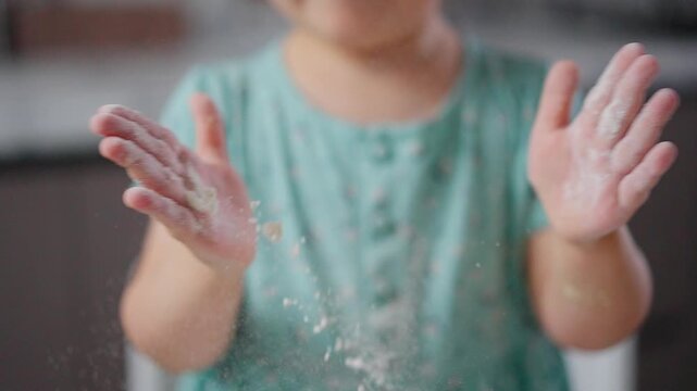Child wears light green dress with floral pattern. Hands shake loose flour in slow-motion. Joyful child laughs while flour dusts air. Perfect for playful kitchen or cooking scenes