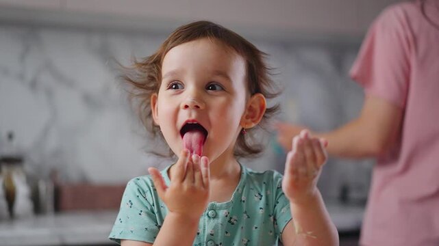 Toddler smiles while tasting a pink substance. Child's hands are raised, showing excitement. Kitchen backdrop features marble countertops. Footage captures joyful, curious exploration