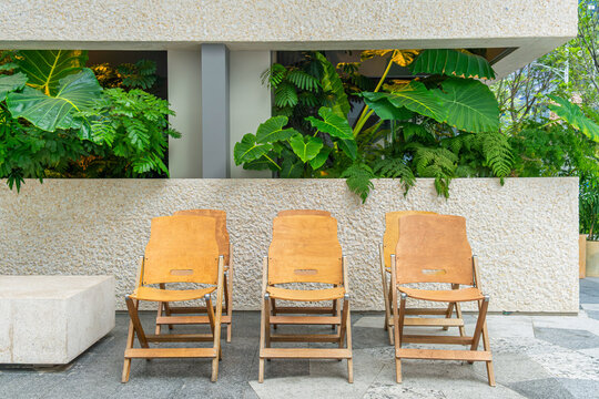 Wooden folding chairs stand in symmetrical row against stone wall and lush tropical plants in Shenzhen city, China. Outdoor furniture mockup, empty seating template, tropical interior design