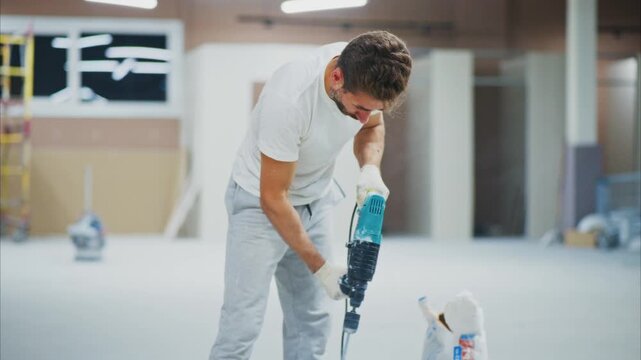 Worker mixes concrete with electric drill in a construction site. Hands steady grip holds tool during mixing process. Scene shows active construction, work in progress. Ideal for construction