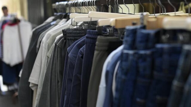 Various garments hang neatly on hangers along clothing rails in a store interior. Additional clothing racks and blurred background activity remain visible during the display shot.