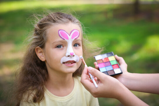 Woman drawing bunny on little girl's face outdoors, closeup