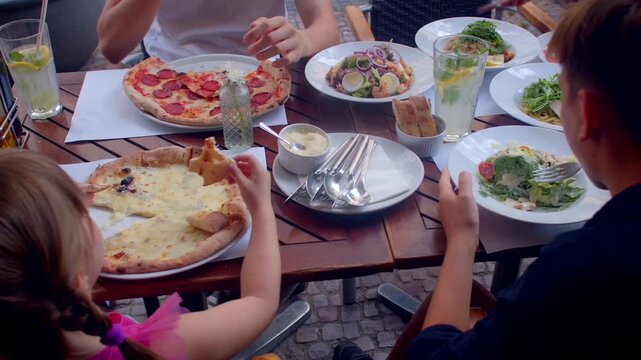 Family dining together and enjoying Italian cuisine. Diners seated on outdoor table pull pizza slices, eat salad. Family members enjoying Italian cuisine and spending time together around the table.