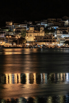 Illuminated harbor and coastal town at night in Kalymnos Island, Greece. Scenic view of Orthodox church and traditional waterfront buildings reflecting on calm sea.