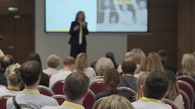 Business conference audience seated in rows, listening to speaker and presentation on stage. Corporate training, networking and professional development atmosphere.