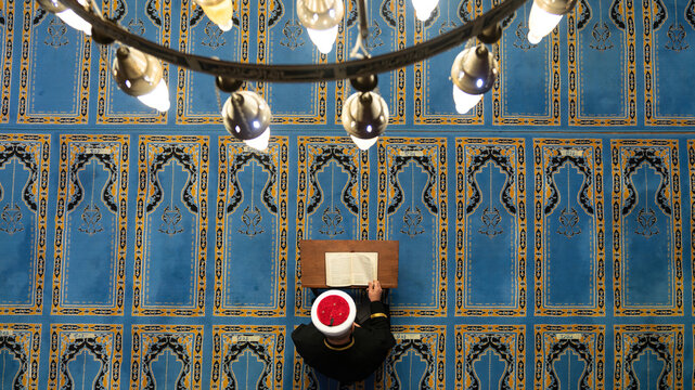 Mosque interior in Savsat, Artvin, Turkey, seen from above during Ramadan: imam in a white turban reading the Quran at a wooden lectern on blue prayer rug carpet