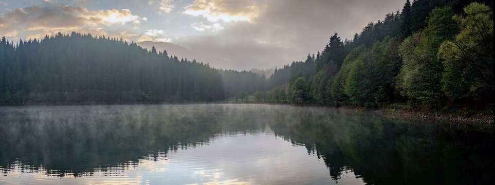 Savsat Karagol lake in Artvin, Turkey, with morning mist rising over the still water, fir forest on the shores and soft light through the clouds, panoramic view
