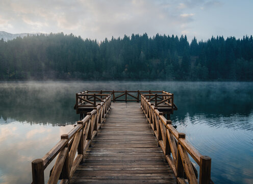 Wooden pier reaching into Savsat Karagol lake in Artvin, Turkey, with morning mist over the water and a dense fir forest on the far shore