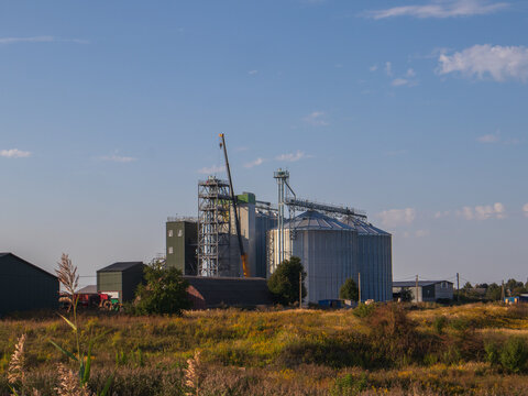 A factory is seen in a rural area with several silos and a crane working. The setting is under a clear blue sky during the late afternoon. Fields surround the site