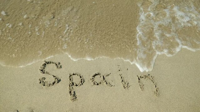 Gentle ocean wave washing away spain word written on sand. Clear ocean water with white foam washing away the word spain written by hand on a sandy beach, representing the end of a vacation