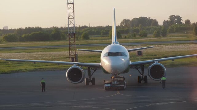 Narrow body passenger airplane with tow tractor attached at stand, ground crew preparing for pushback, warm sunset light