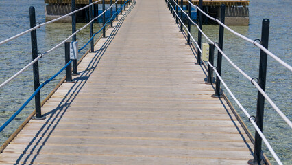 Naklejka premium Long wooden pier stretches over clear shallow tropical water with black metal railings and white rope barriers on both sides under bright sunny sky during day in coastal vacation setting.