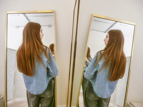 Young woman stands between two mirrors, facing away. Long brown hair flows down her back. She wears a denim shirt and dark jeans. Calm and confident, she checks her outfit. Perfect for fashion, beauty