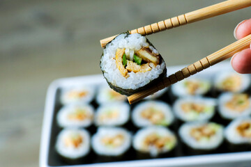 Maki sushi chopsticks holding. Home made Japanese maki sushi rolls with rice, cucumber and tofu chicken. Fresh rolled sushi close up food photography. Horizontal photo of Asian chopstick usage.  © Laura