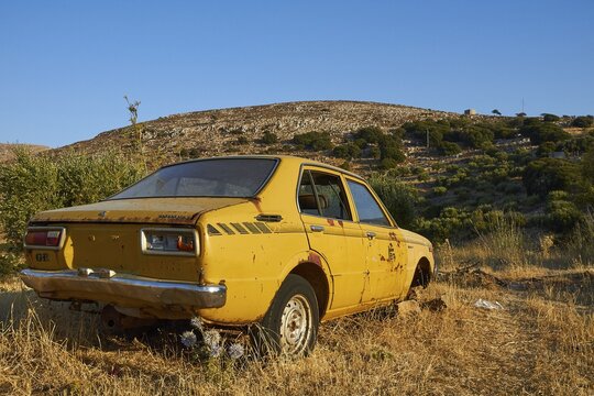 Yellow rusted car in a dry landscape surrounded by greenery under a sunny sky, wrecked vehicle, Crete, Greek Islands, Greece