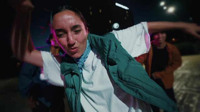 A woman in a green vest and white shirt dances toward the camera in an outdoor Barcelona plaza lit by magenta and purple LED lights at night.