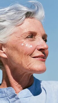 Happy senior woman with silver hair and sunscreen dots on her cheek enjoying the sun against a clear blue sky, evoking SPF skincare and joyful aging