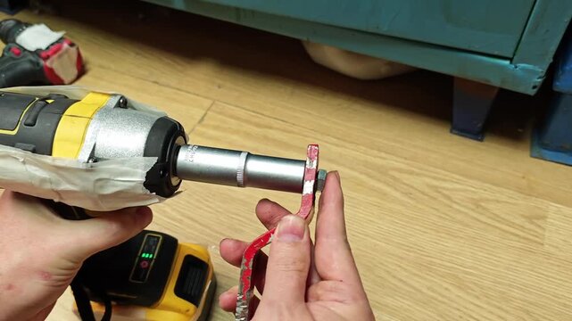 Hands position hex key beside impact driver during tool setup. Driver extends fastener while fingers hold red tool steady on wood floor, aligning socket extension for precise manual adjustment.