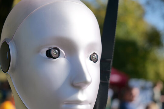 White humanoid robot head with camera lens eyes, glossy plastic android face closeup, artificial intelligence cyborg with neutral expression, blurred sunny outdoor background