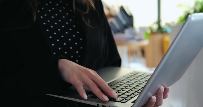 Professional female employee types messages on wireless laptop while standing indoors. Determined worker manages digital project tasks using portable computer within modern company workplace.