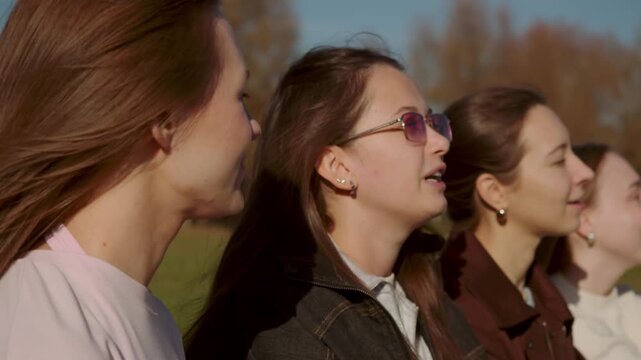 Four young female friends chat and smile outdoors in autumn park. Close up side view captures happy women sitting in row under warm sunlight and enjoying peaceful weekend nature.