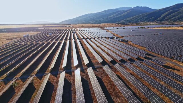 Rows of solar panels are aligned on flat land in a remote area. The setting shows a bright sky and green mountains. The scene is warm and bright.