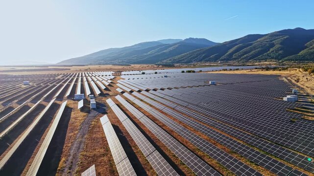 A large field covered with solar panels captures sunlight. The location is in a mountainous region with a clear blue sky during daytime.