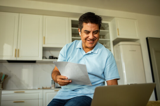 Smiling man reviewing document while using laptop in kitchen