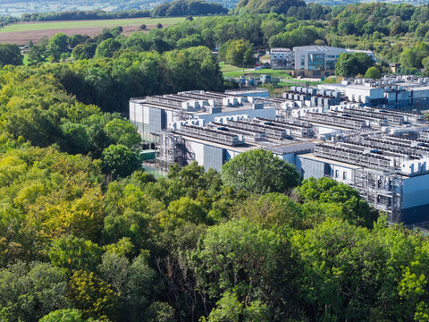 Aerial view of a modern data center facility nestled within a dense forest, featuring industrial cooling units on the roof and surrounding green trees in the United Kingdom.