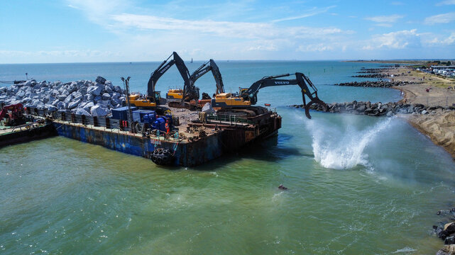 Aerial view of two excavators on a barge unloading large rocks into the sea for coastal defense in Jaywick, Essex, United Kingdom.