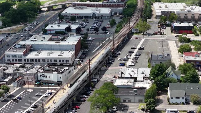 Parallel railway tracks divide commercial buildings and parking areas in a high angle suburban drone view, showing the rail line and surrounding streets from above.