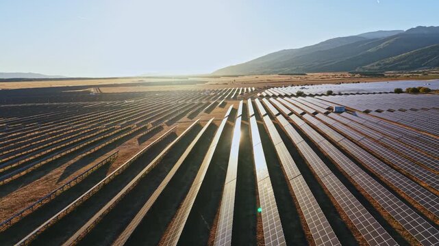Solar panels cover a large area on the slopes of a hill. The sun sets in the background, casting light across the rows of panels. The location appears to be open land.