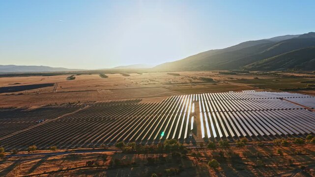 Rows of solar panels stretch across a vast field in a rural area. The sun rises over the mountains casting light on the landscape.