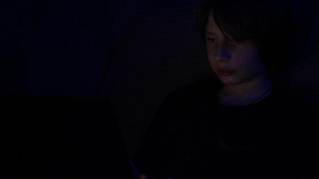 A young boy sits in a dark room late at night, deeply focused on his laptop screen. The glowing light illuminates his face as he browses the internet or plays video games