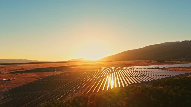 The sun rises over a solar farm with multiple rows of solar panels. The landscape shows open fields and distant mountains under a bright sky.