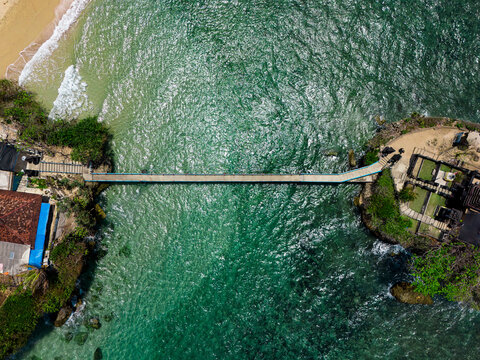 Aerial view of the bridge connecting the rocky shore to the temple island at Balekambang Beach, surrounded by clear turquoise water and coastal vegetation in Malang, East Java, Indonesia.