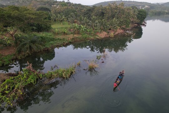 Aerial view of a fisherman in a wooden canoe on the calm Volta River, with lush tropical vegetation and palm trees along the riverbank in Akosombo, Eastern Region, Ghana.
