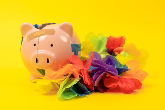 A pink piggy bank mended with tape and a bandage rests next to a bright tropical flower garland on a yellow background, a concept of vacation spending and holiday travel costs.