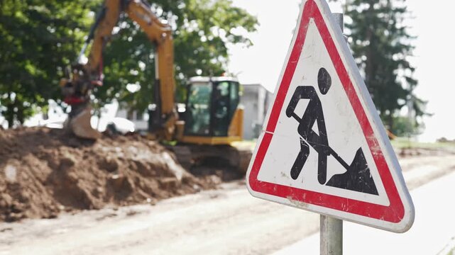 Construction area features a warning sign for work in progress, with an excavator digging earth and trees visible in the background under bright sunlight