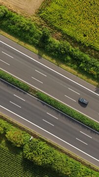 Vertical screen. Aerial perspective captures two vehicles traveling on a highway bordered by lush greenery and agricultural fields, illustrating the serene countryside