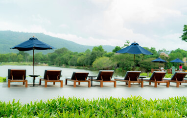 Fototapeta premium Luxury resort poolside with wooden lounge chairs and blue umbrellas beside a tranquil lake and green mountain during daytime, offering a tropical vacation retreat for relaxation and sustainable travel