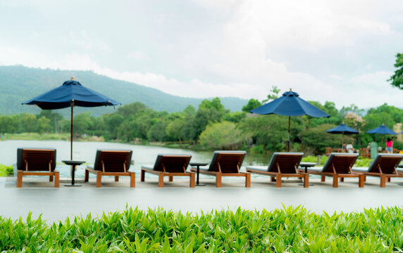 Luxury resort poolside with wooden lounge chairs and blue umbrellas beside a tranquil lake and green mountain during daytime, offering a tropical vacation retreat for relaxation and sustainable travel