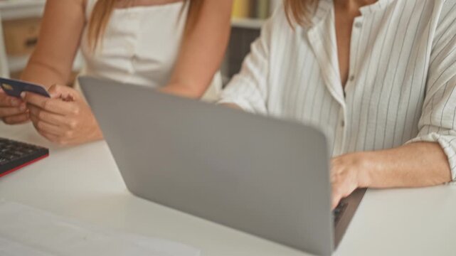 Woman and older woman lean over laptop, one holds creditcard and points to screen in a building; focused collaboration.
