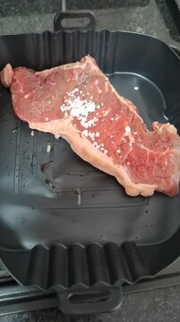 Close-up of a raw cut of beef being seasoned with coarse salt before cooking, showing a simple food preparation step on a dark air fryer tray