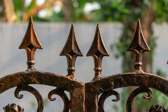 wrought iron fence. Rusted iron spikes stand tall and sharp. Curved metalwork frames the pointed tips. Weathered surface hints at age and exposure. Faded greenery blurs softly in the background