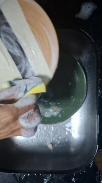 Person scrubs a plate with a sponge and dish soap in a stainless steel kitchen sink, showing manual dishwashing and household cleaning after a meal
