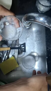 Close-up of hands rinsing drinking glasses under running water in a stainless steel kitchen sink, capturing a simple household dishwashing routine