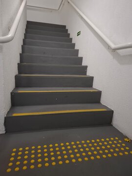 Empty indoor staircase in an administrative building with dark gray steps, yellow anti-slip edge markings, tactile warning paving and white handrails for safer pedestrian access