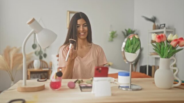 Woman applying makeup brush to cheek at vanity table with mirror and smartphone in studio; self care confidence.