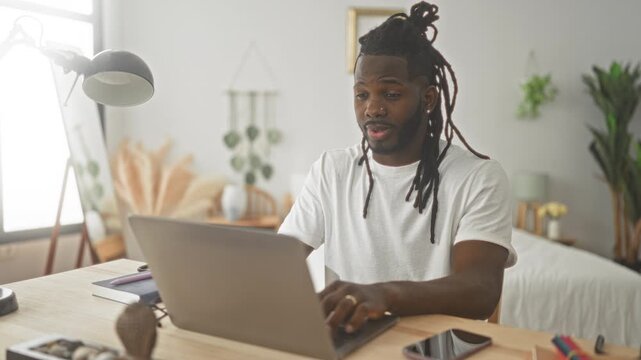 Man points finger to camera and types on laptop at tidy bedroom desk, smiling and engaging; playful remote work connection.